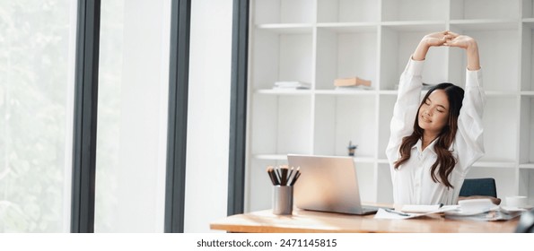 A young woman stretches at her desk in a modern office, taking a relaxing break from work. The bright workspace features large windows and shelves in the background. - Powered by Shutterstock