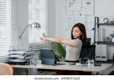 A young woman stretches at her desk in a modern home office, surrounded by office supplies and a digital tablet, promoting a healthy work environment. - Powered by Shutterstock