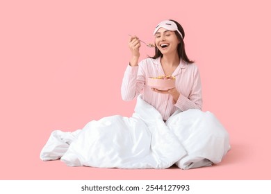 Young woman in striped pajamas with blanket eating cereal rings on pink background - Powered by Shutterstock