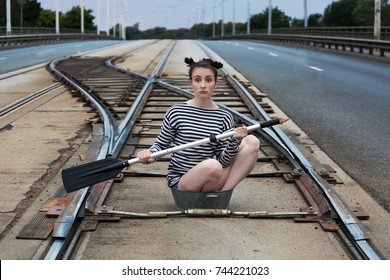 Young Woman In A Striped Sailor's Vest And With Oar In Hands In The Middle Of Tram Rails Sits In Tin Little Basin - Strange Place And Device For Navigation 