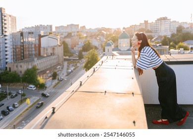 Young woman in striped shirt and red shoes leans on rooftop terrace overlooking cityscape at sunset. Urban buildings in background. . Concepts of freedom, contemplation, tranquility, reflection - Powered by Shutterstock