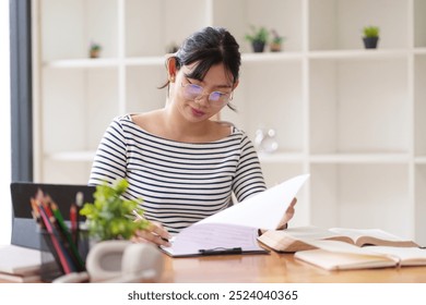 A young woman in a striped shirt is sitting at a desk, looking at a piece of paper while working on her laptop. She is in a bright room with large windows, surrounded by natural light. - Powered by Shutterstock