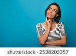 A young woman in a striped shirt stands against a blue background with a thoughtful expression, resting her finger on her chin. Her pose suggests curiosity or contemplation.