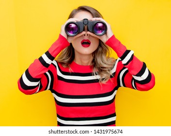 young woman in striped sweater with binocular on yellow background 