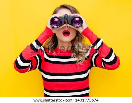 young woman in striped sweater with binocular on yellow background 