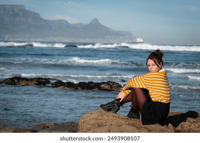 A young woman in a striped sweater sitting on rocks by the sea with mountains in the background - Powered by Shutterstock