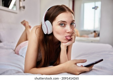 Young woman stuck out her tongue while lying in bed in wireless headphones with phone in her hands.