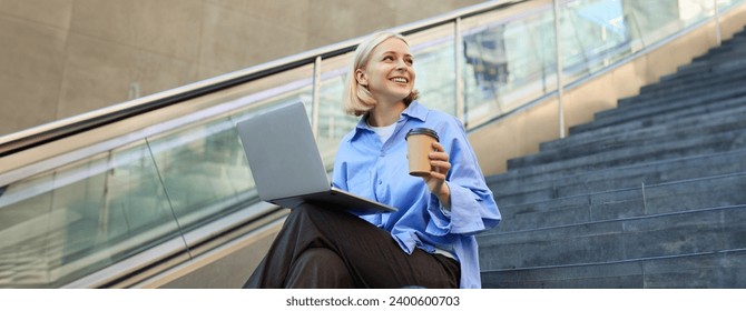 Young woman, student in blue shirt, working on laptop, sitting outdoors on street stairs, working on project online, connects to public wifi, elearning. - Powered by Shutterstock