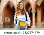 A young woman student with curly hair holds several colorful notebooks while standing outside a historic university building. University Student On Campus