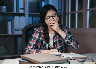 Young Woman Student At Home Desk Reading And Yawning Tired At Night With Pile Of Books. Exhausted College Girl Preparing Exam In Midnight Stay Up Late Hard Working. University Education Concept