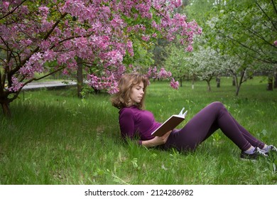 young woman, student, reads book with interest, lying under tree on spring day in park. girls - teenager 17 years old with book on grass. preparation for exams. Spring leisure. Passion for reading