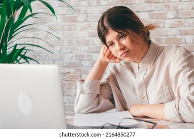 Young Woman Student Sitting At Desk Full With Books Textbooks Looking At Computer Screen To Unfinished Homework Feels Tired Bored And Unmotivated, Girl Loaded With Tasks, Work Backlog Overflow Concept