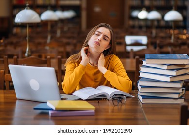 . A Young Woman Student Is Sitting At A Table Full Of Books With Textbooks, Feeling Tired, She Is Massaging Her Neck, Back Pain. Girl Loaded With Tasks, Work Backlog Overflow Concept