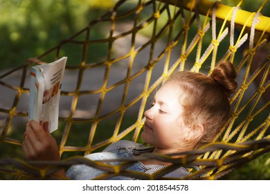 Young Woman Student Smiling Reading A Book While Lying On A Hammock In The Garden. Woman Relaxing In Hammock Reading Book. Back To School, University, Campus.                              