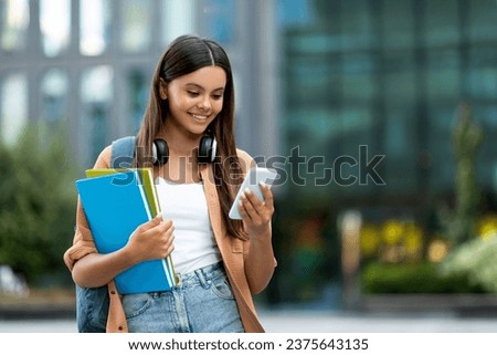 Young woman student texting on smartphone at campus, copy space. Positive cheerful lady with backpack and notebooks in her hand walking by street, using phone, copy space. Communication