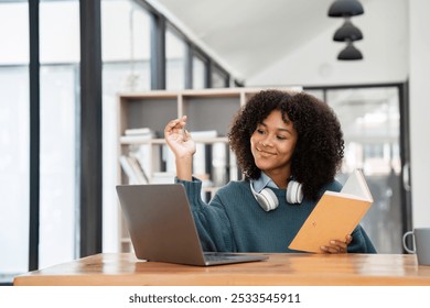 Young Woman Studying Online at Home with Laptop and Book, Wearing Headphones, in Modern Bright Room - Powered by Shutterstock