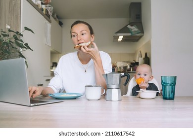 Young Woman Studying Or Working Online At Home While Having Breakfast With Her Baby In Kitchen. Millennial Mother On Maternity Leave With Child. Freelancer Busy Mom With Laptop Searching Information.