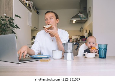 Young Woman Studying Or Working Online At Home While Having Breakfast With Her Baby In Kitchen. Millennial Mother On Maternity Leave With Child. Freelancer Busy Mom With Laptop Searching Information.