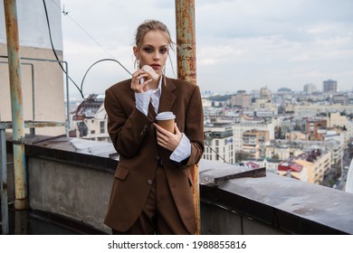 young woman in stylish outfit holding paper cup and donut on rooftop with cityscape on blurred background