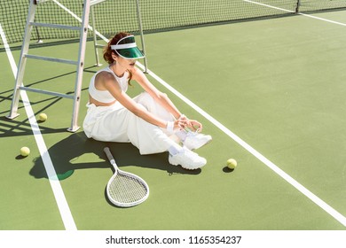 young woman in stylish white clothing and cap tying shoelaces on tennis court with racket and balls - Powered by Shutterstock