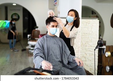 Young Woman Stylist Wearing Face Mask And Gloves Working In Hairsalon Doing Haircut And Hair Styling For Male Client, New Normal Due To Coronavirus Outbreak