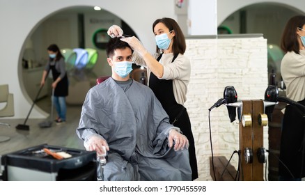 Young Woman Stylist Wearing Face Mask And Gloves Working In Hairsalon Doing Haircut And Hair Styling For Male Client, New Normal Due To Coronavirus Outbreak