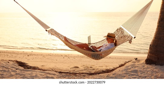 Young Woman, Successful Female Freelancer Using Laptop While Lying In Hammock On The Tropical Beach At Sunset, Working Remotely During Summer Vacation. Distance Work Concept