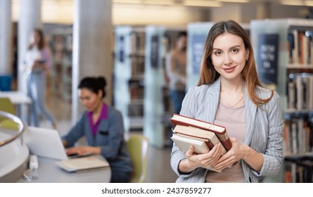 Young woman successful student standing with books in library, other students choosing books and working on background behind - Powered by Shutterstock