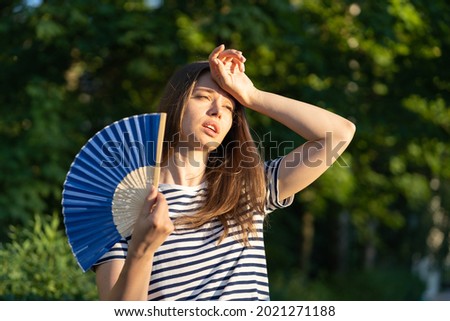 Young woman suffering heatstroke outdoors using fan to get fresh air. Unhappy girl feel bad of hot temperature outside. Frustrated female tired of summer season heat in fatigue touch forehead sweating