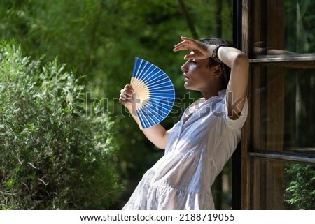 Young woman suffering heatstroke outdoors using fan to get fresh air. Unhappy girl feel bad of hot temperature outside. Frustrated female tired of summer season heat in fatigue touch forehead