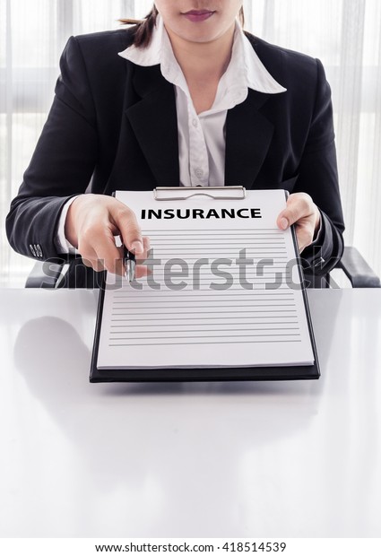 young woman in suit in his office showing an\
insurance policy and pointing with a pen where the policyholder\
must to sign