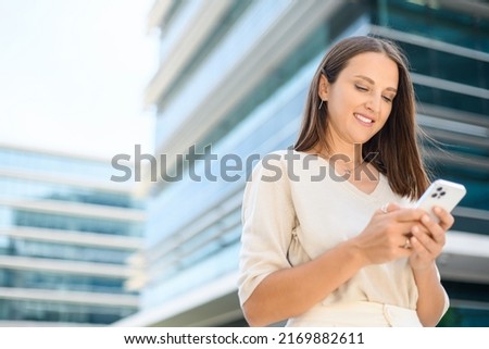 Young woman suit stands on the city street in downtown and using smartphone. Carefree female office employee using cellphone on city street with urban background, enjoys online chating