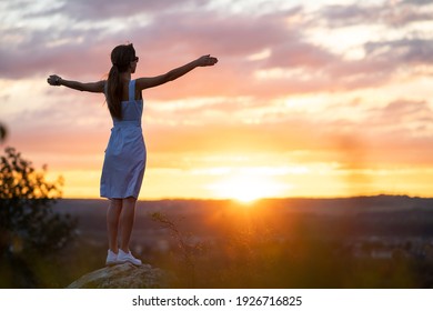 A young woman in summer dress raising up her hands standing outdoors enjoying view of bright yellow sunset.