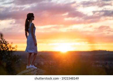 Eine junge Frau im Sommerkleid steht draußen und genießt den Blick auf hellgelben Sonnenuntergang.