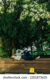 A Young Woman In Summer Lies On A Wooden Bench Under A Large Tree In The Shade