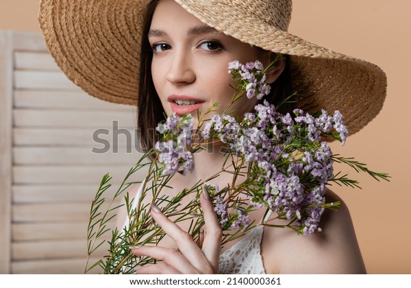 Young woman in sun hat\
holding flowers and looking at camera near blurred folding screen\
isolated on beige