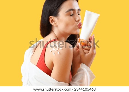 Young woman with sun made of sunscreen cream on her shoulder blowing kiss against yellow background, closeup