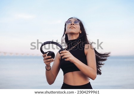 Young woman in sunglasses and black headphones dancing and smiling, listening to the music by the river side, sunset light 