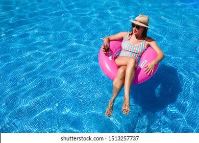 Young Woman With Sunglasses, Hat And Swimsuit In A Blue Pool. Pretty Girl On A Pink Float Enjoying The Summer While Having A Cocktail.