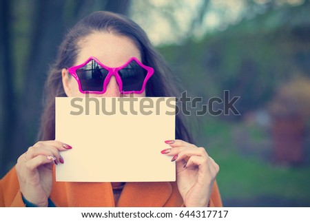 Young woman in sunglasses holding blank sheet of paper near face