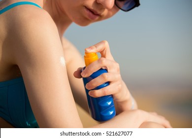 Young woman in sunglasses holding bottle of sunscreen lotion, spraying sunblock cream on shoulder before tanning, close up. Sunburn and cancer prevention concept