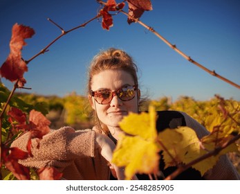 A young woman with sunglasses smiles gently at the camera from within a vineyard. Golden and red autumn leaves frame her face. The sun shines brightly in a clear blue sky. - Powered by Shutterstock