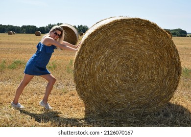 A young woman in sunglasses smiling jokingly pushes a round haystack in a large cereal field - Powered by Shutterstock