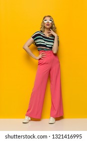 Young woman in sunglasses and striped blouse and pink wide legs trousers is posing with hand on hip. Full length studio shot on yellow background. - Shutterstock ID 1321416995