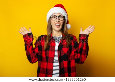 Young woman with surprised face and santa hat and red checkered shirt on yellow background