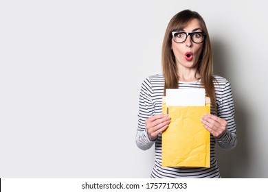 Young woman with a surprised face takes out a letter or notice from a paper envelope on a light background. Concept letter from college, taxes, ticket