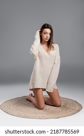 Young Woman In Sweater And With Bare Legs Kneeling On Carpet On Grey Background