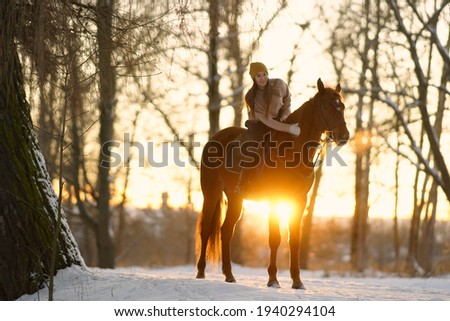 Young woman in sweater and cap riding a brown horse on snow at sunset. Background of winter trees and sunset sky