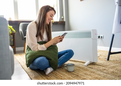 Young Woman In Sweater Feeling Cold, Waiting For The House To Heat Up, Controlling Heating System With A Smart Home Thermostat. Woman And Radiator Using Smartphone App For Temperature Control