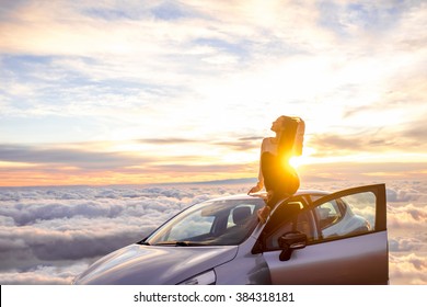 Young Woman In Sweater With Heart Shape Enjoying Beautiful Cloudscape Sitting On The Car Roof Above The Clouds On The Sunrise. Wide Angle Image With A Lot Of Space